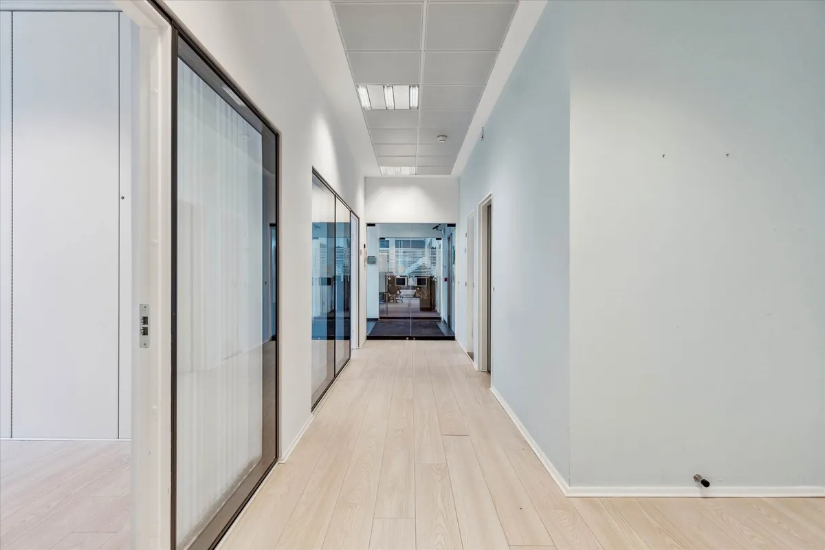 Lang og smal gang med lysegul trægulv og hvide vægge samt høje, glasede døre til rum med vinduer og møbler