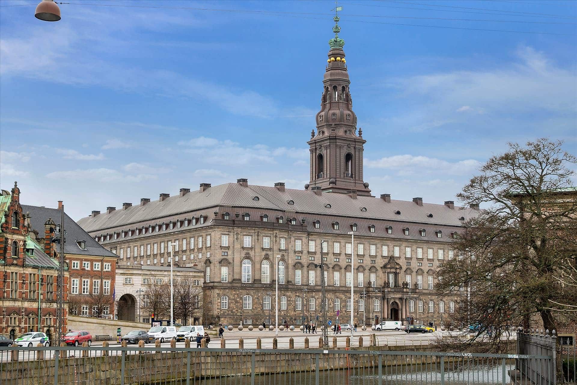 Københavns middelalderby med bygning med spire og en række parkerede biler foran bygningen og en person går i baggrunden