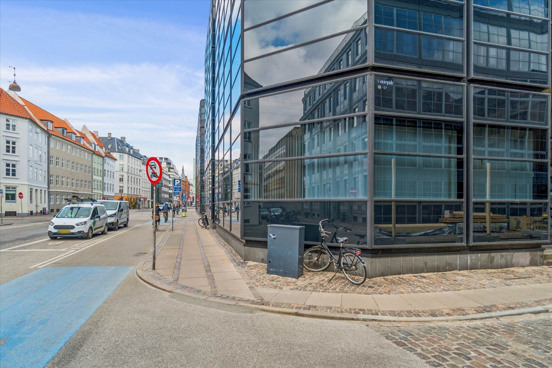 Moderne bygning med glasfasade og parkerede cykler. Strømlinet vej med to biler og cykelstier. En cykelparkeringsboks står