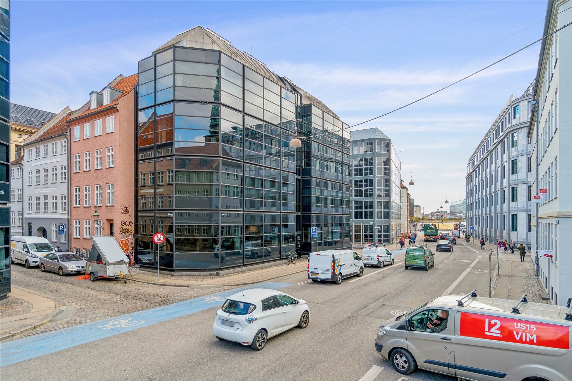 Gadekorsning med biler og folk i baggrunden og en stor bygning med glas og refleksioner i fronten