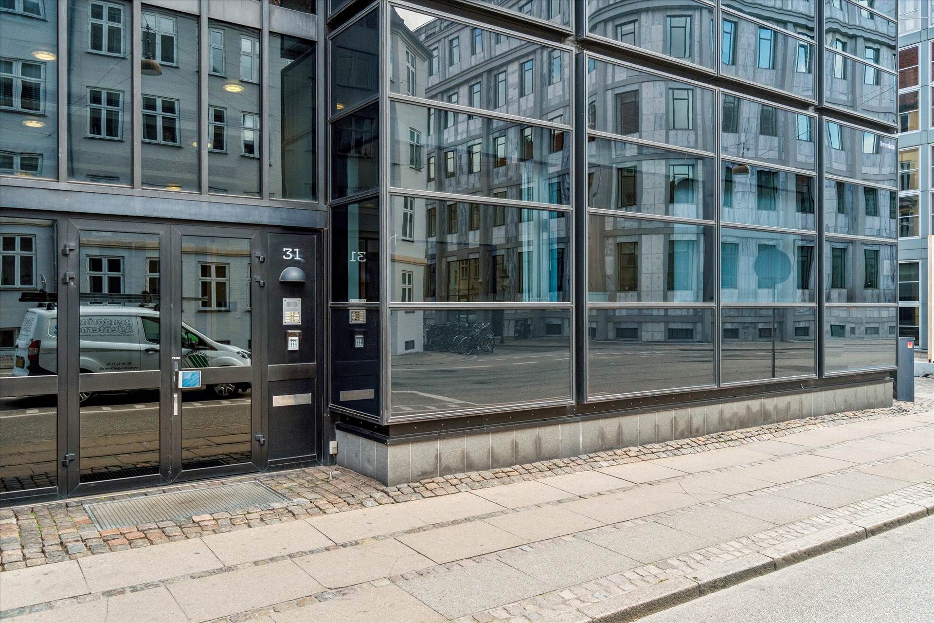 Markant ejendom med glasvinduer og nummer 31, foran hvilken er parkeret en hvid van. På gaden er der flere cykler, og