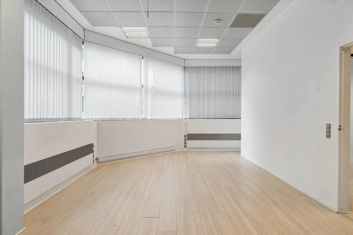 Lyst og stort kontor med højloft og lyseruder. Lys trægulv og hvide vægge med store ruder. Stående radiatorer og hvide