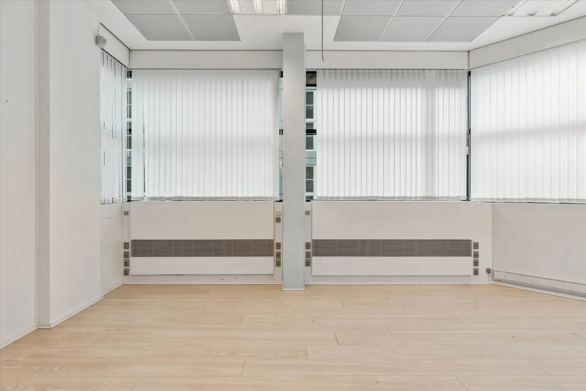 Et stort, lyserum med højt loft, vinduer og hvide jalousier, gulv med træpaneler og radiatorer under vinduer