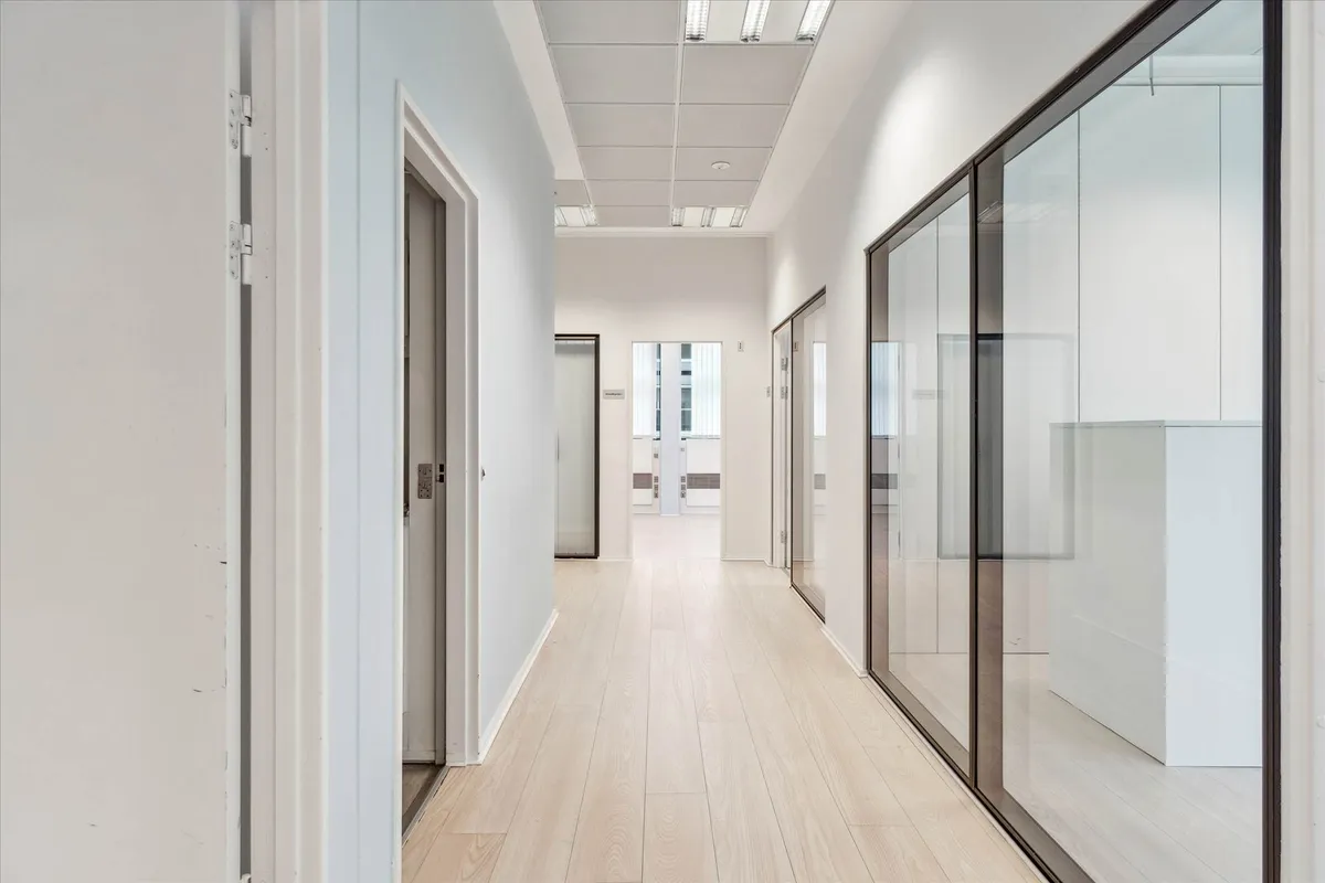 En lys og rummelig korridor med hvide vægge, glasvinduer og lyse trægulve. Stærkt fokus på moderne og minimalistisk design