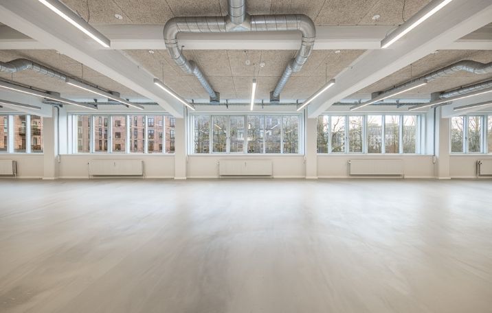 Et stort, lyst og tomt kontor med hvide vægge, højt til loftet og store vinduer med udsigt til bygninger og træer