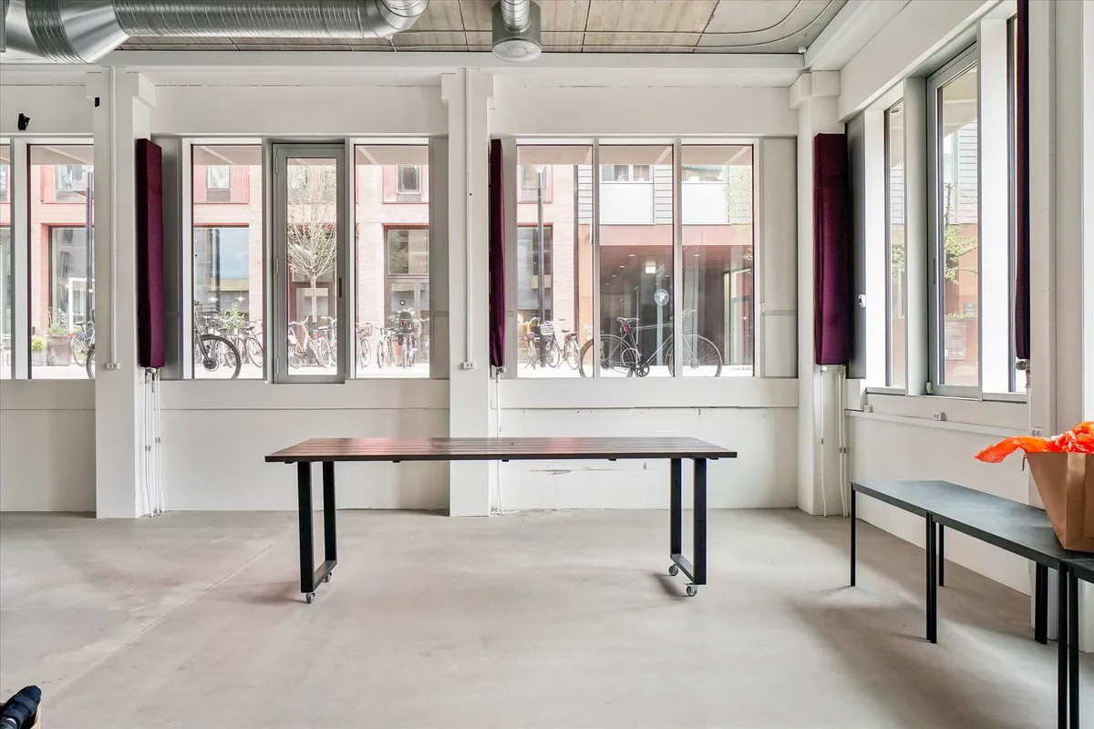 En stor tom rum med en lang bord og to bænke samt vinduer med mørke gardiner og udsigt til bygninger og cykler i baggrunden