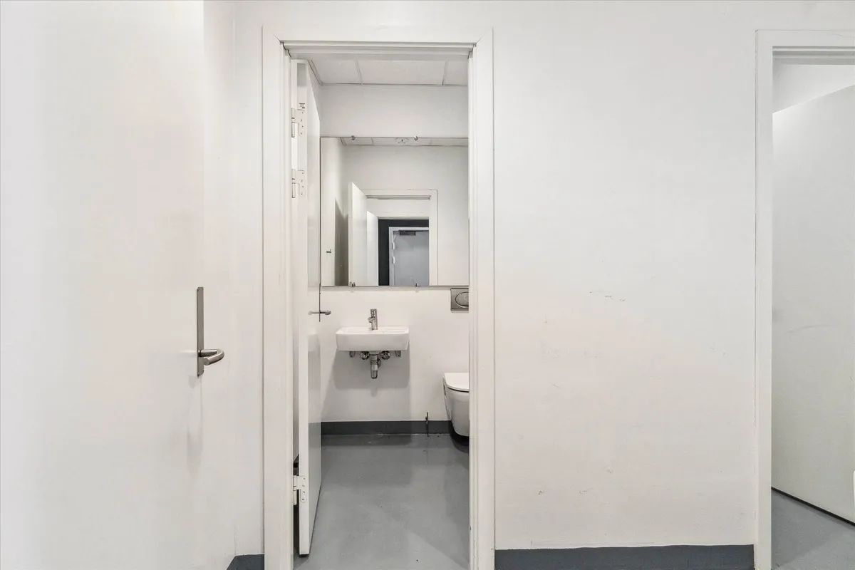 Et rent, minimalistisk designet badeværelse med toilet, håndvask og spejl, der reflekterer dørens åbning.