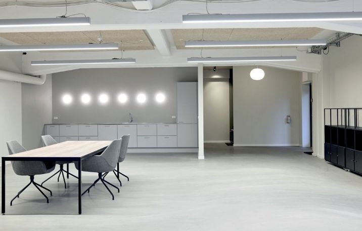 Kontor med langt bord og fire stole langs væggen, høje loftslamper, lysegrå vægge og hvidgulv