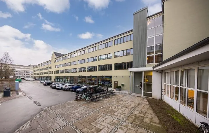 Kontorbyggeri med parkeringsplads og biler, facaden er lysegul og glasvinduer, en række bænke og plantekasser med grønne