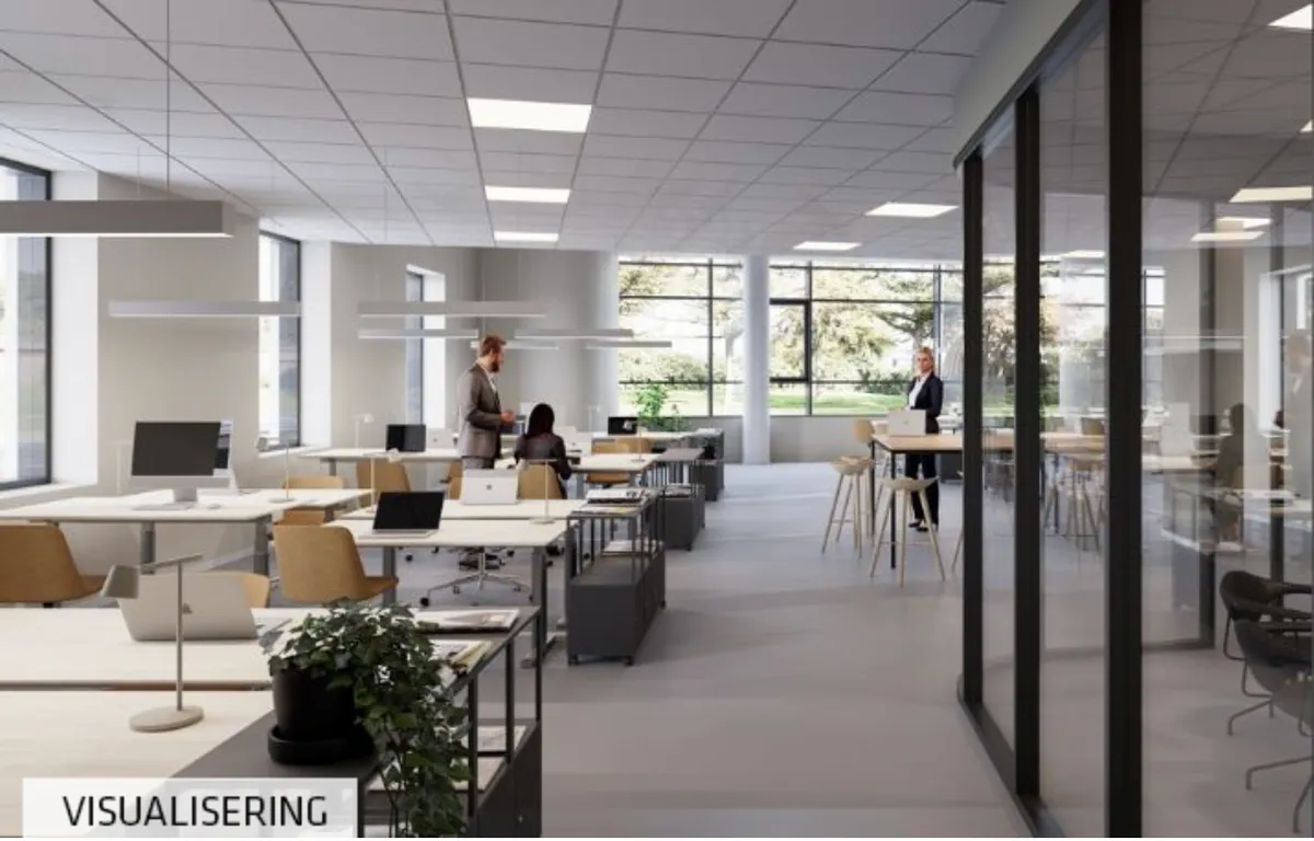 Moderne kontor med lyse loftsvinduer og højt gulv. Folk arbejder ved computere, og der er lyse gulve og store glasvinduer