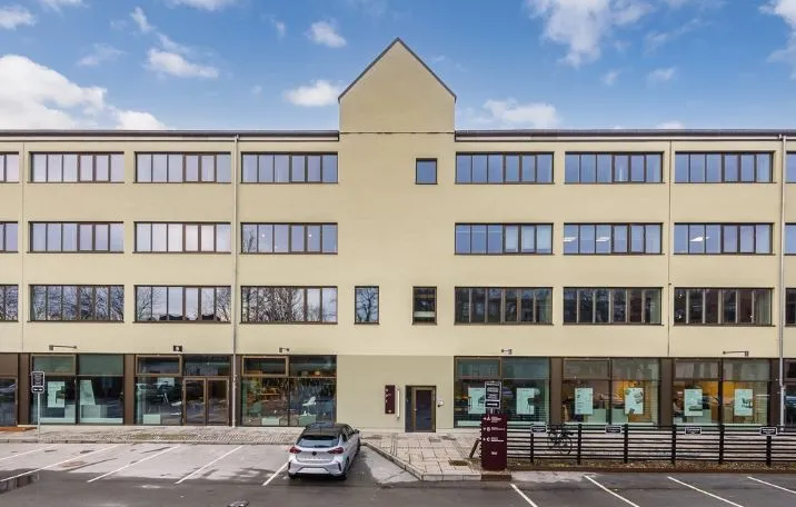 Kontorby i lysegult udenfor med store vinduer og en parkeringsplads med en bil foran.