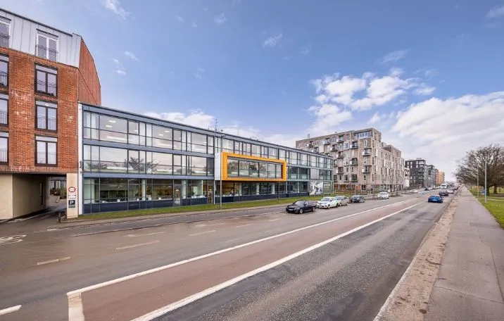 Kontorbyggeri med glasfacade og gul accent på en travl gade med biler, bygninger og tre i baggrunden