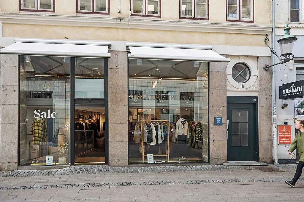 En butik med udsalgsskilte, hvor en kvinde går forbi med en grøn jakke og sorte bukser og en buskeplante i potte