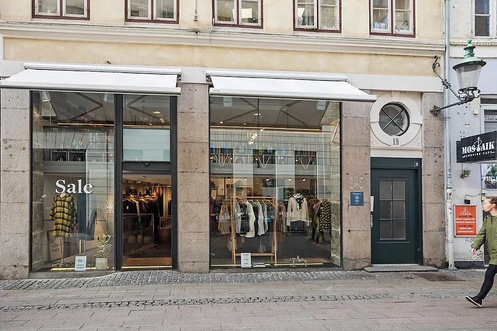 En butik med udsalgsskilte, hvor en kvinde går forbi med en grøn jakke og sorte bukser og en buskeplante i potte