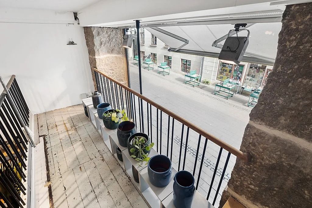 En terrasse med flere potter og en grøn plante samt udsigt til et tomt gadeforløb med bord og stole