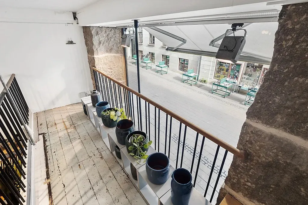 En terrasse med flere potter og en grøn plante samt udsigt til et tomt gadeforløb med bord og stole