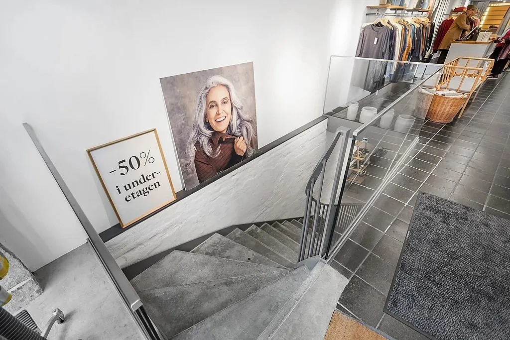 Trætrapper og udstillingsvindue med plakater og kurve i en forretning. En kvinde ser ud af et vindue, og der er tøj og en