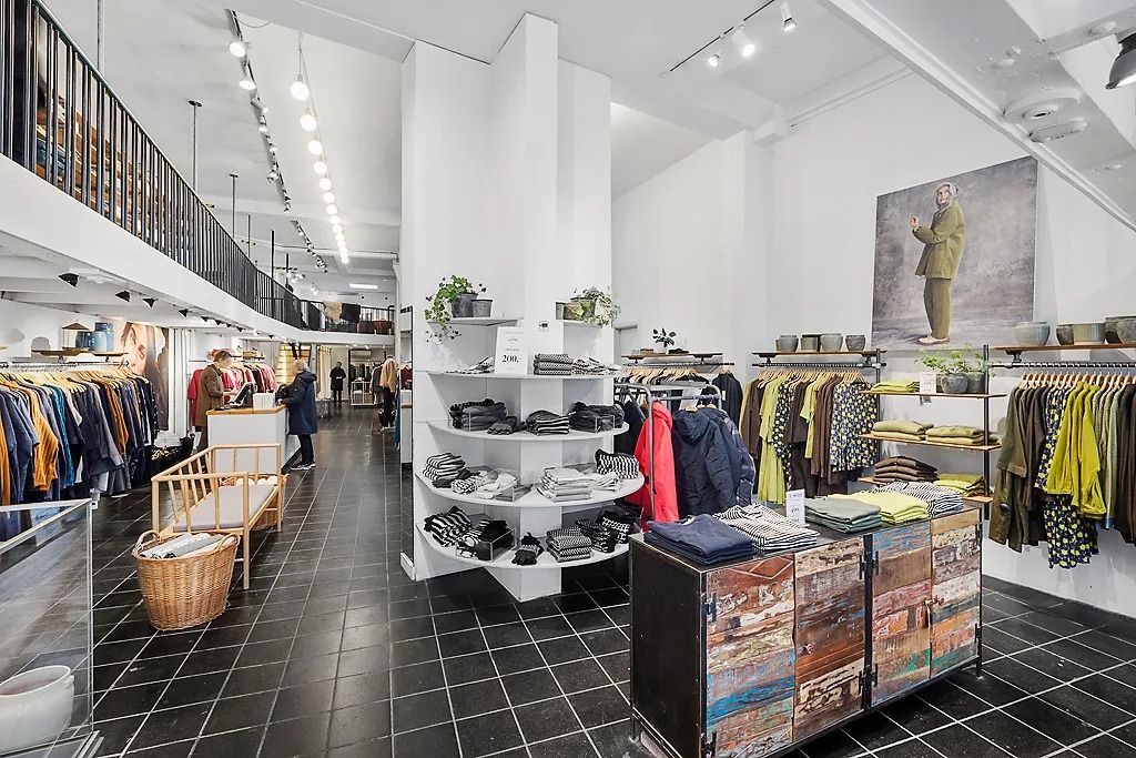 En stor tøjbutik med lyse loftslamper og plantepoter på hylderne. Sort og hvidt tøj og sjaler er placeret på en høj hylde.