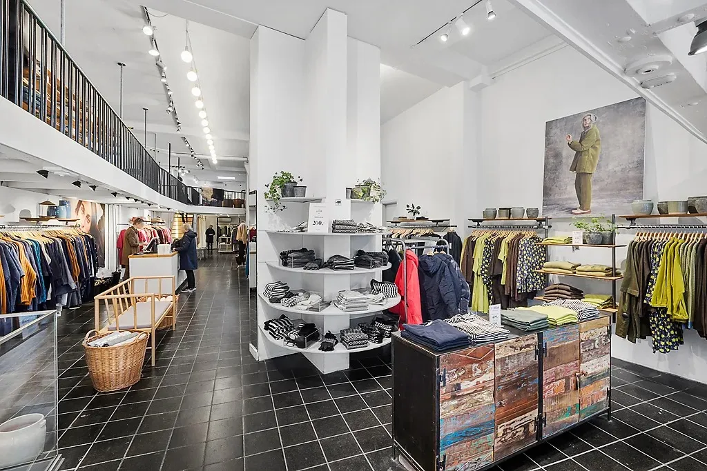 En stor tøjbutik med lyse loftslamper og plantepoter på hylderne. Sort og hvidt tøj og sjaler er placeret på en høj hylde.