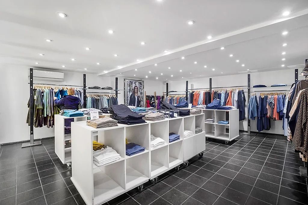 Indvendigt i en moderne, veludstyret tøjbutik er der flere høje, hvide hylder med tøj, der er opdelt i kategorier. Der er en