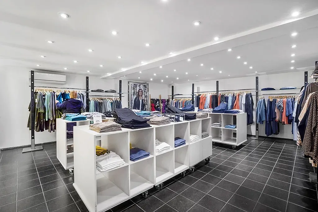 Indvendigt i en moderne, veludstyret tøjbutik er der flere høje, hvide hylder med tøj, der er opdelt i kategorier. Der er en