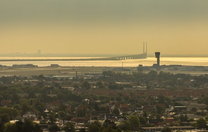 Luftfoto af Københavns Lufthavn med bro, tårn, bygninger og hav, set i dagslys med lyserøde himmelfarver og smukke