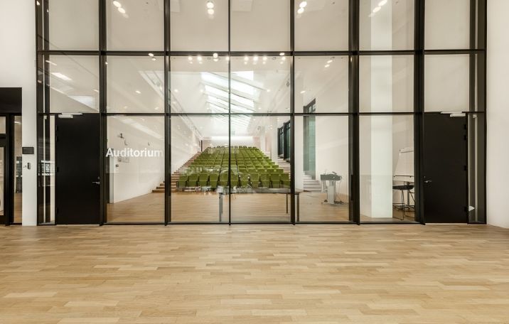 Glass- og trædøre med auditorium-skilt, to stolpe med skrivebord, gulve og vægge i lyse nuancer, og en række grønne stole i