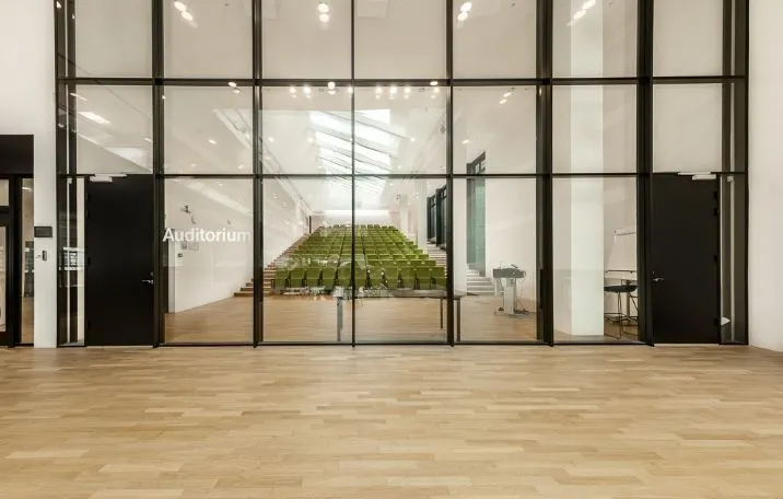 Glass- og trædøre med auditorium-skilt, to stolpe med skrivebord, gulve og vægge i lyse nuancer, og en række grønne stole i