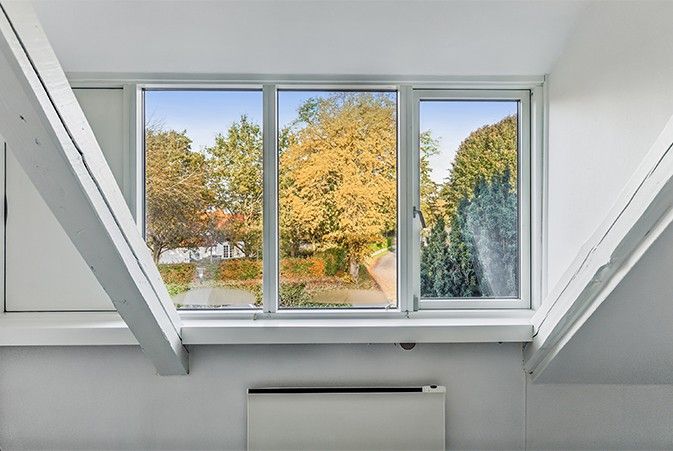 Vindue med udsigt til lyse guldfarvede træer og en hvid hus i baggrunden. Vinduet er lukket og er udstyret med hvide rammer