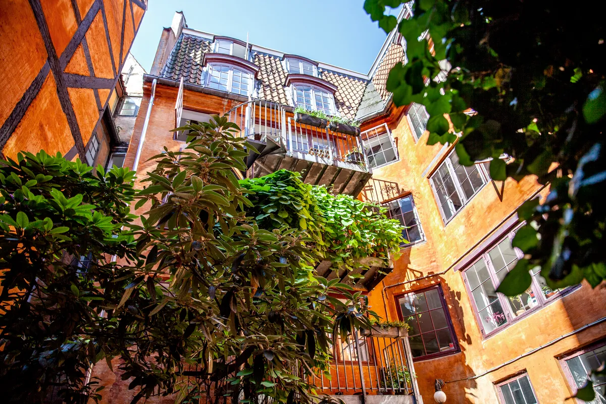 En gammel bygning med terrasse og plantepot, som er renoveret og malet i orange. Der er træplanter foran bygningen og en