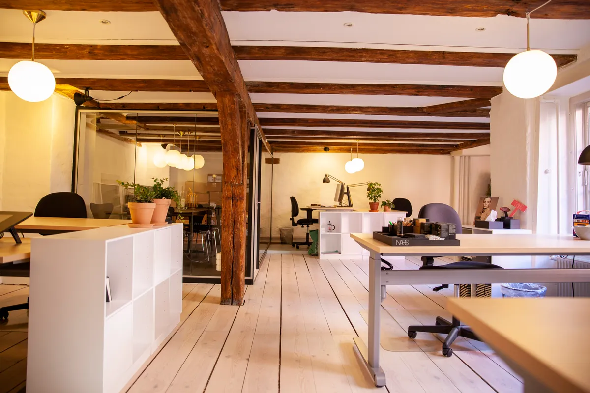 Kontor med højt loft og lyse loftsbjælker, gulve og vægge samt moderne møbler og plantager i lyse potter