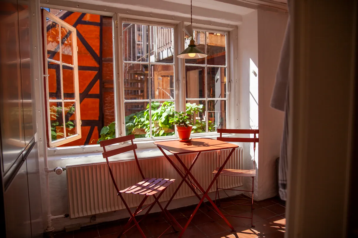 Smukt udstyret lille køkken med røde stole, træbord og plante. På bordet står en lysekroneslampe, og i vinduet er der en