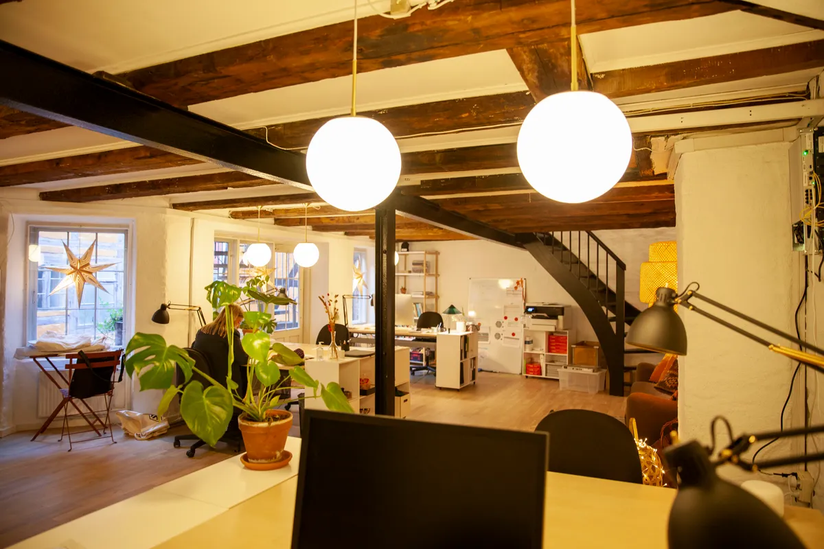 Kontor med lyse gulvbelægning, tre store runde lysekroner, borde, skriveborde og en computer. Der er en sofa og et bord med