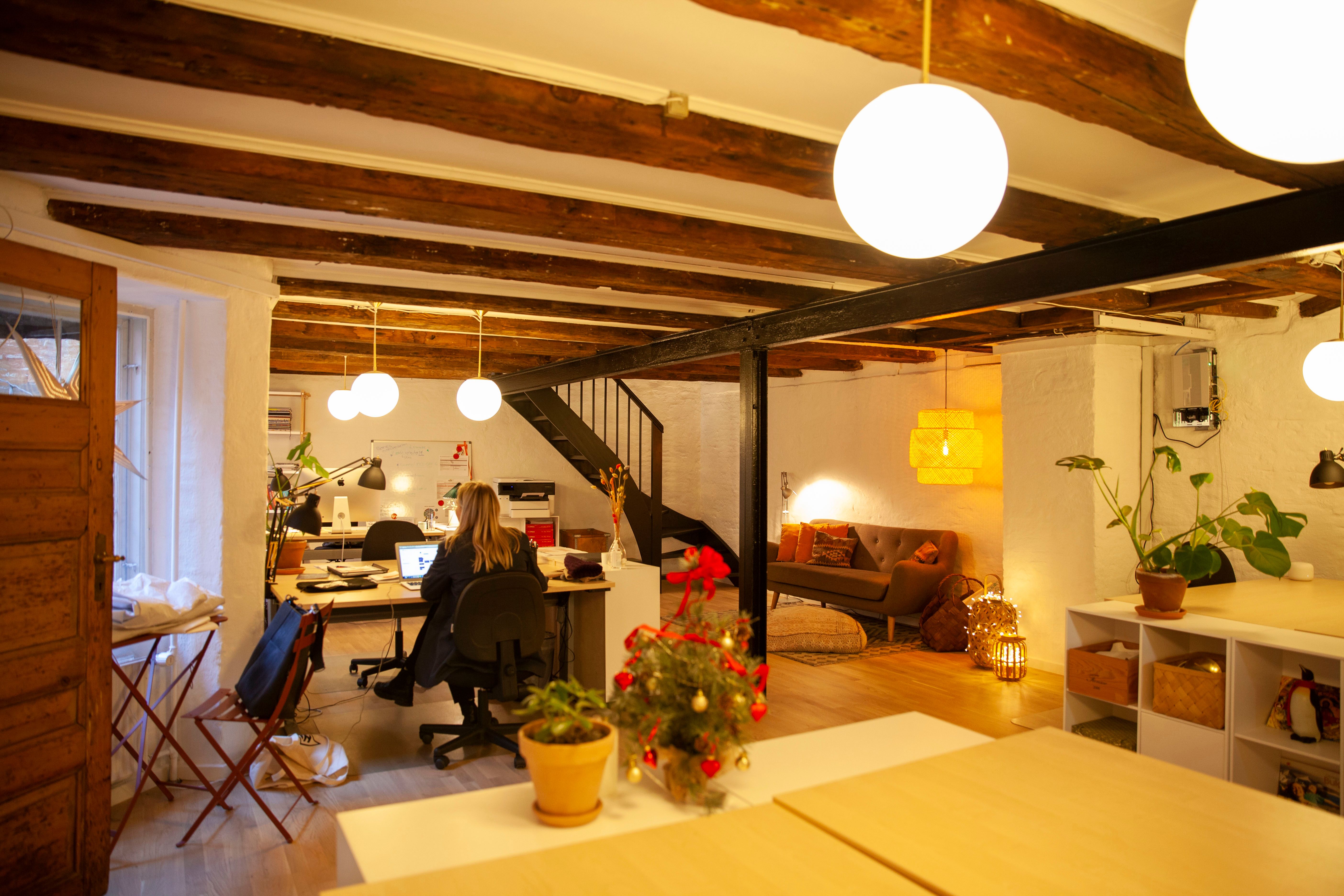 Kontor med lyse loftsbjælke og lysekroner, to personer arbejder i kontoret, sofaer og plante i stuen, gulv med mønster