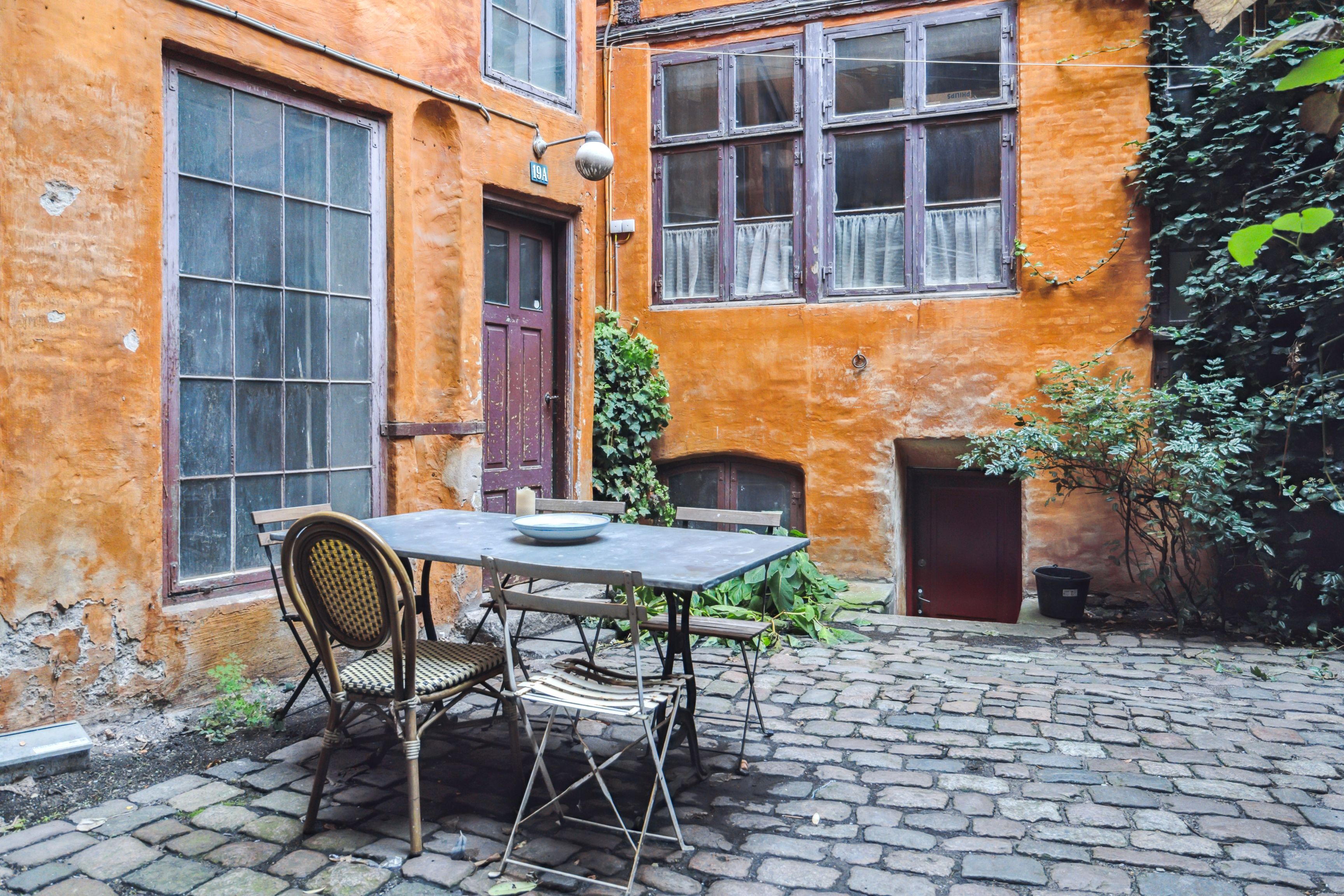 Møbelstang med fire stole og bord i gårdhave med gammelt gule bygningsfacade og vækst af planter og buske