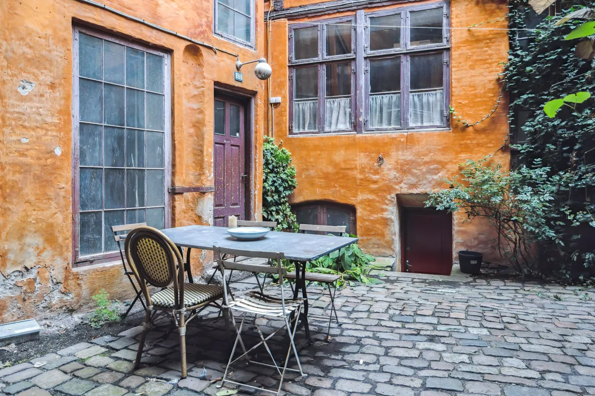 Møbelstang med fire stole og bord i gårdhave med gammelt gule bygningsfacade og vækst af planter og buske