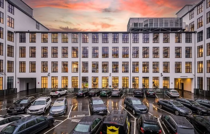 En moderne kontorbygning med sandblæste søjler og glasvægge i sorte sprosse parkeret med biler i regnvejr med skyerne i