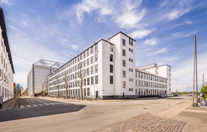 Stort hvidt kontorhus med glasvægge og sandblæste søjler i byen, med parkerede biler og cykler foran