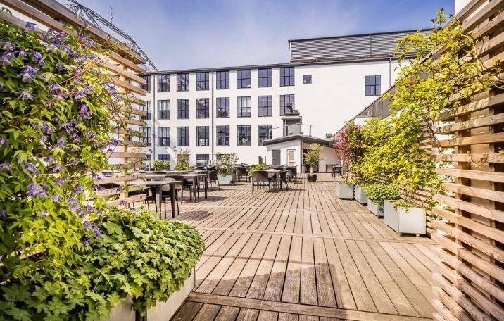 Træterrasse med borde og stole i skygge og planter i potte. Solskin trænger igennem og kaster skygger. Bygning i baggrunden