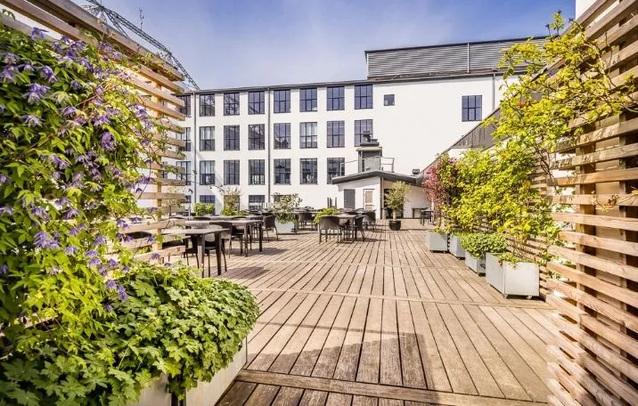 Træterrasse med borde og stole i skygge og planter i potte. Solskin trænger igennem og kaster skygger. Bygning i baggrunden