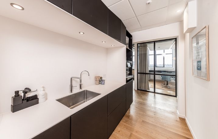 Kontor med rustfrit stål køkken, hvidt bord, sort køkkenskab, lysekroner og åbent glasdør til mødelokale