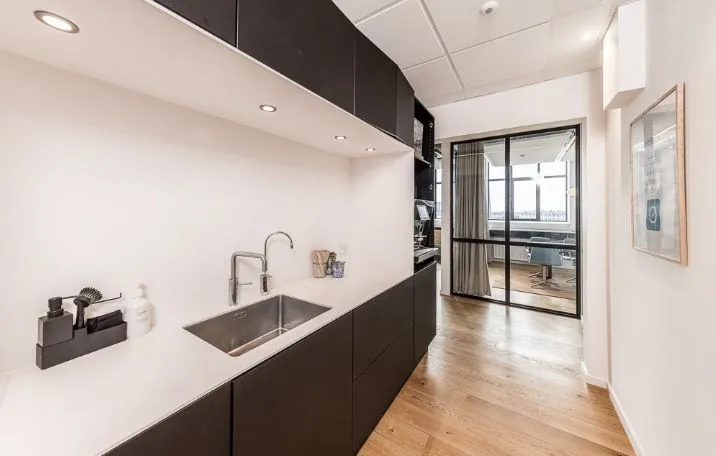 Kontor med rustfrit stål køkken, hvidt bord, sort køkkenskab, lysekroner og åbent glasdør til mødelokale