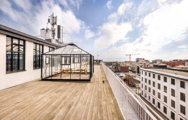 Lyst kontorlokal i rå fabriksstil med trægulv, glasvægge i sorte sprosser, søjler og et glasdækket område med skovlhøjder