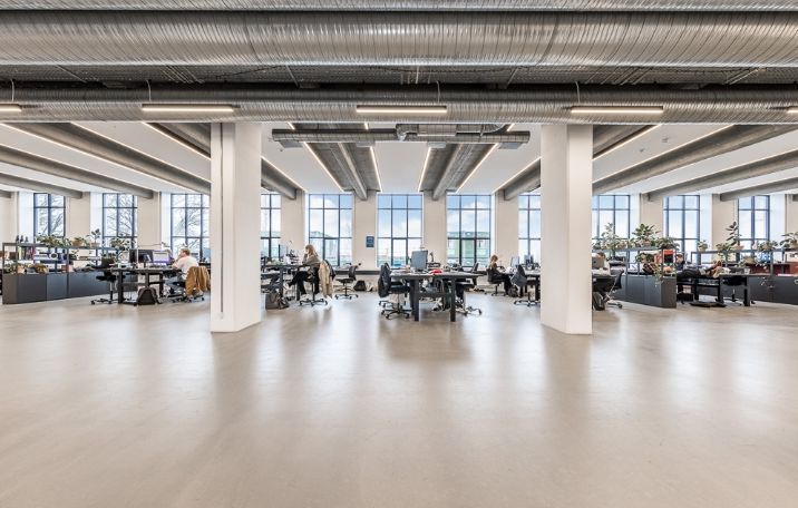 Kontorrum med store glasruder og højloft. Arbejdsborde og stole er placeret langs væggene. Værelset er møbleret med