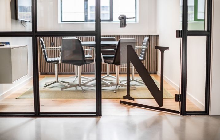 Moderne mødelokale med lyse gulve og store glasvægge, der viser bogstavet N i sort og lysegrå. Hjørnebord og stole med