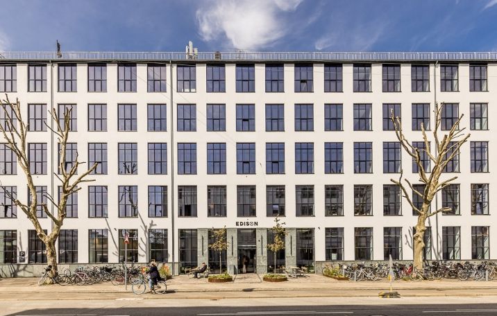 Stort kontorbygger i rå fabrikstil med Edison på facaden og mange glasvinduer, cykler parkeret foran og to træer i forgrunden