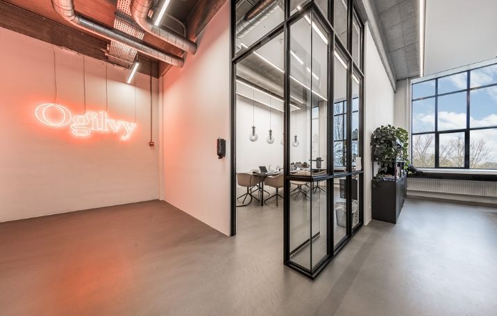 Moderne kontor med åbent rum, sandblæste søjler, glasvægge og neon-logoet Ogilvy på væggen