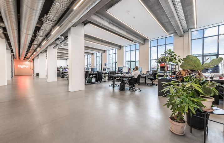 Kontor med åbent areal, sandblæste søjler, glasvinduer, bord med computer og plante, arbejdspladser og lyse bælter på loftet