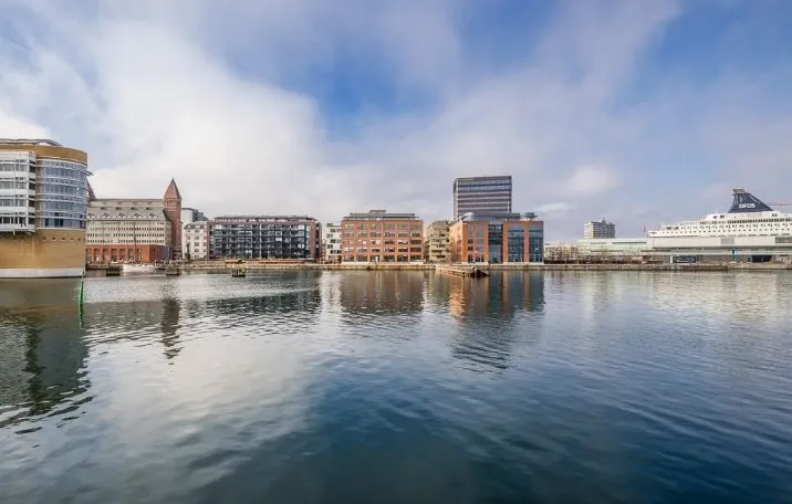 Byens skyline med havn og skib med tårn, flere højhuse og bygninger i baggrunden, refleksioner af bygninger og skib i vandet