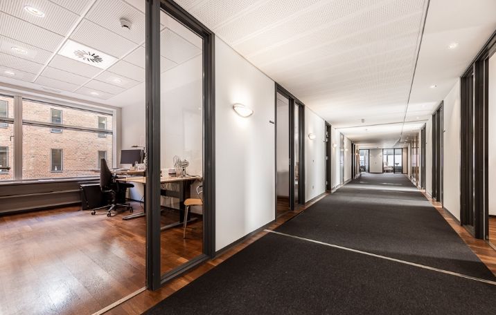 Moderne kontorlokaler med glasvægge og lysegule lofter. Væggene er hvide og der er en skrivebord med computer.